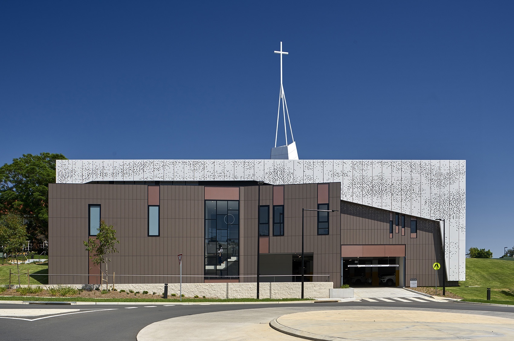 Südseite der St. James Chapel mit modernen dunklen Keramikpaneelen und einer oberen Struktur aus perforierten Metallplatten, die von einem großen Kreuz gekrönt wird, umgeben von einer gepflegten Grünfläche und einem Gehweg