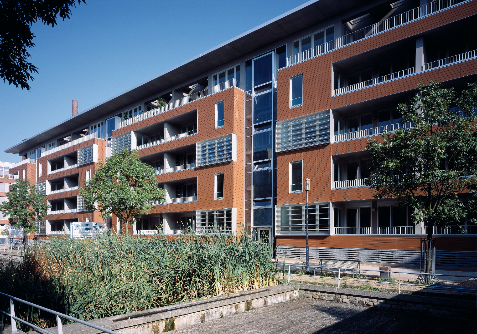 Frontansicht eines Wohnungsbauprojekts im Innenhafen Duisburg mit roten Keramikpaneelen, modernen Balkonen und großen Fenstern, umgeben von grüner Vegetation