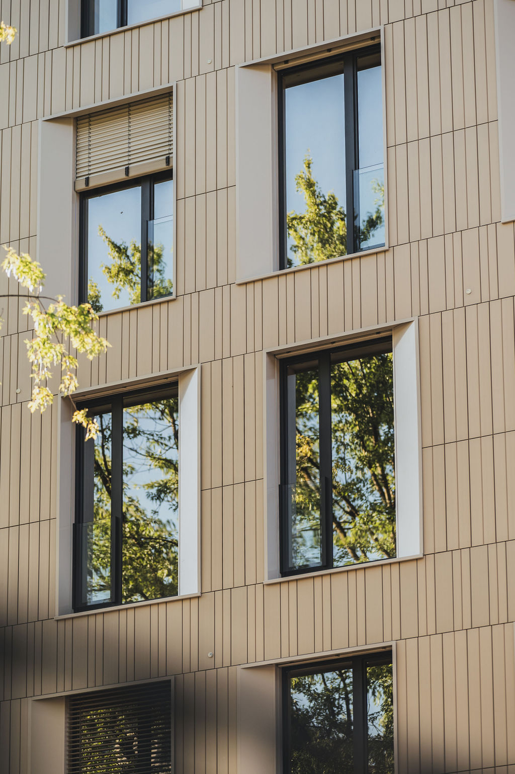 Detailaufnahme der Keramikfassade des Baerahus in Feldkirch mit vertikalen Linien und großen Fenstern, die Bäume reflektieren