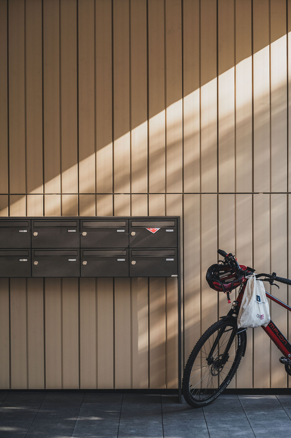 Detailaufnahme der Keramikfassade des Baerahus in Feldkirch mit vertikalen Linien, schwarzen Briefkästen und einem roten Fahrrad im Sonnenlicht