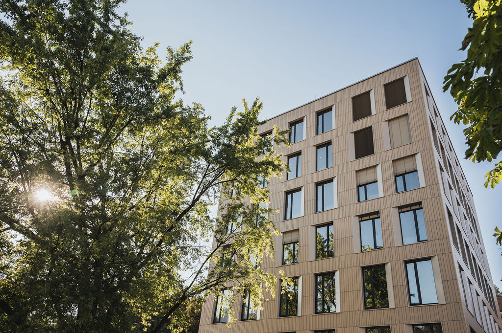 Keramikfassade des Baerahus in Feldkirch mit vertikalen Linien und großen Fenstern, umgeben von Bäumen im Sonnenlicht