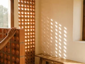 Perforated terracotta ceramic screen panel casting geometric shadows on warm interior wall with golden sunlight filtering through circular openings