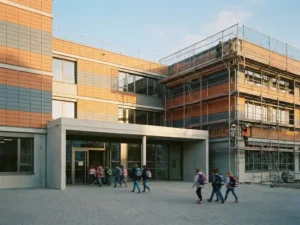 Keramikfassadenplatten in Terrakotta und Schiefergrau werden an einer Schulgebäude-Außenwand installiert, Kinder betreten den Eingang darunter.