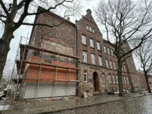 Frisch sanierte Schulfassade mit Terrakotta- und hellgrauen Keramikfliesen, teilweise abgebautem Gerüst und Eichen im Vordergrund.