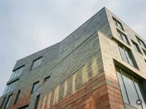 Modernes Gebäude mit Keramikfassade in Terrakotta-, Salbei- und Schiefergrau-Tönen, Nachmittagslicht beleuchtet die strukturierten Fliesen von unten.